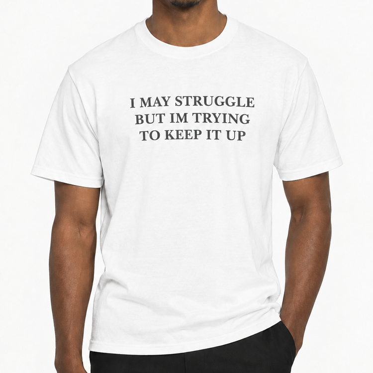 A man wearing a white crew-neck t-shirt with the black printed text 'I MAY STRUGGLE BUT IM TRYING TO KEEP IT UP' on the chest, paired with black pants, against a plain white background.