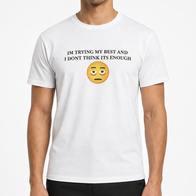A man wearing a white short-sleeve crewneck t-shirt with the text 'I'M TRYING MY BEST AND I DON'T THINK IT'S ENOUGH' above a yellow sad face emoji, paired with black pants, against a light gray background.